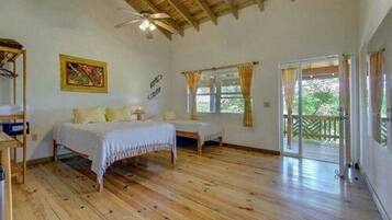 Una habitación con piso de madera, una cama, un escritorio y una silla. Cuenta con una ventana con cortinas y vistas al exterior, con zonas verdes.