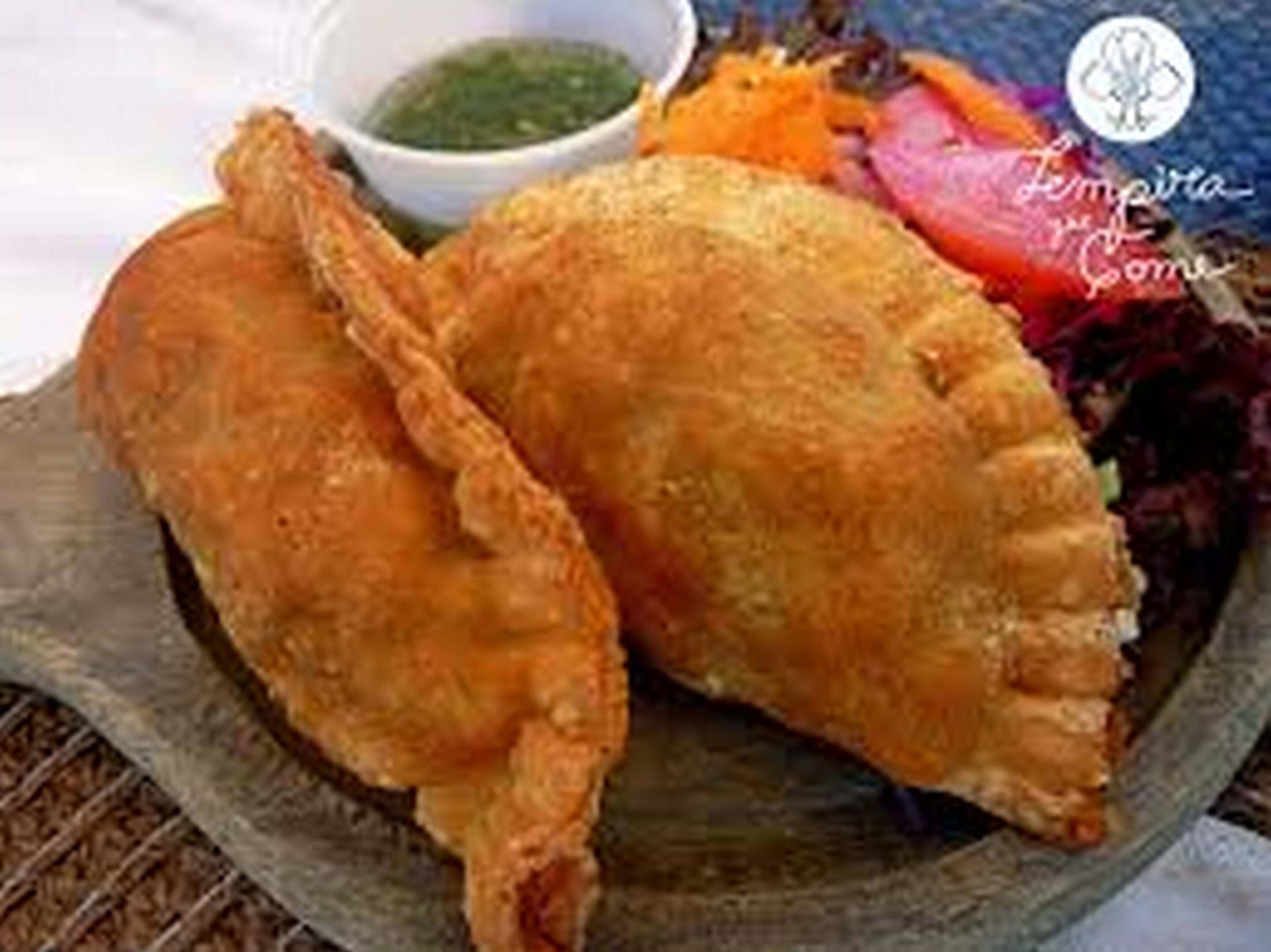 Dos pasteles fritos sobre un plato de madera, acompañados de salsa verde y vegetales mixtos.