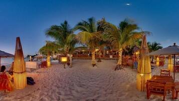 Un resort en la playa con palmeras, mesas y sillas de madera bajo luces colgantes.