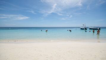 A beach with clear turquoise water, a few people swimming, and boats anchored nearby.