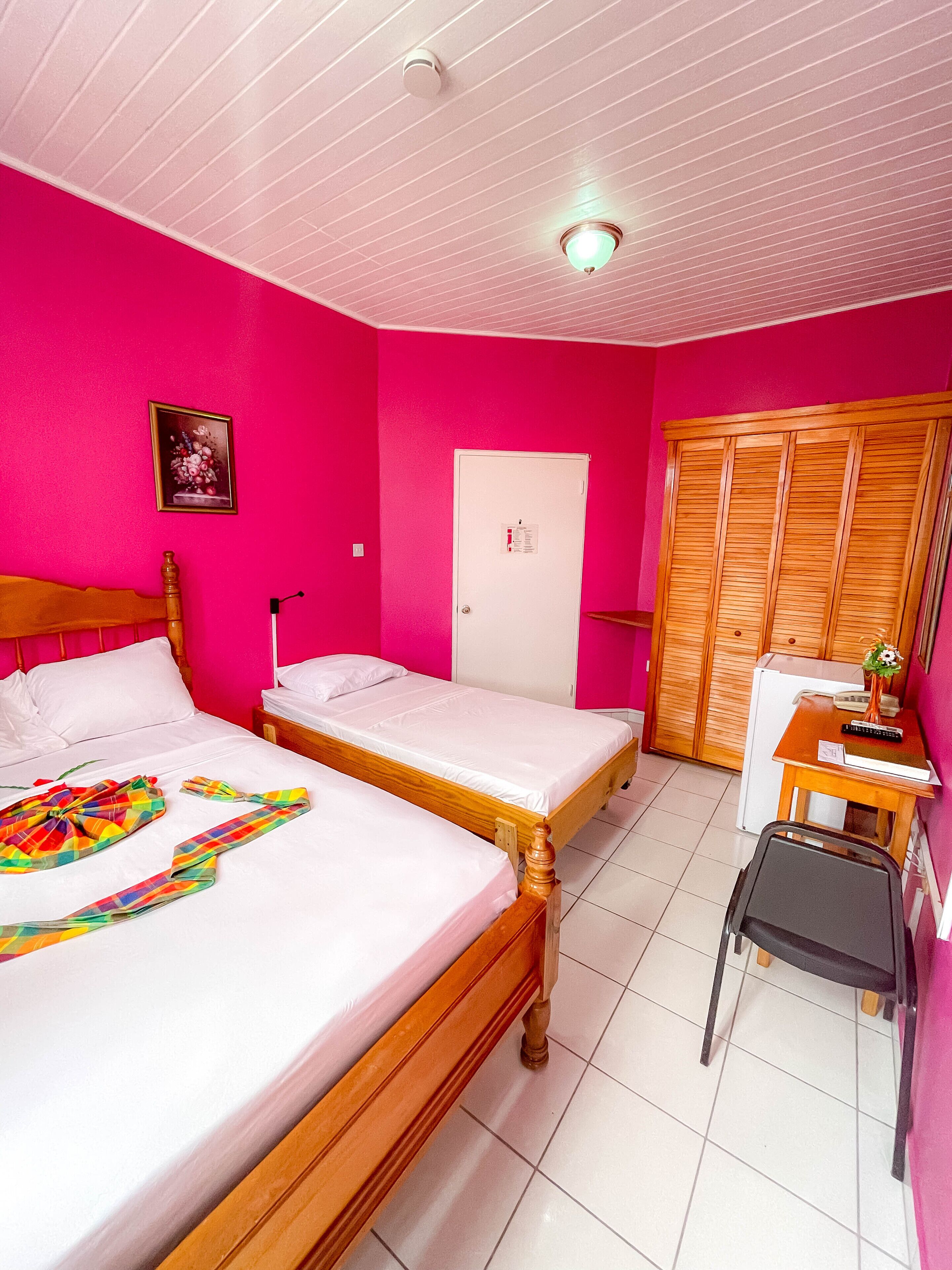 A room with two beds, a wooden wardrobe, a desk with a chair, and a door with a sign.