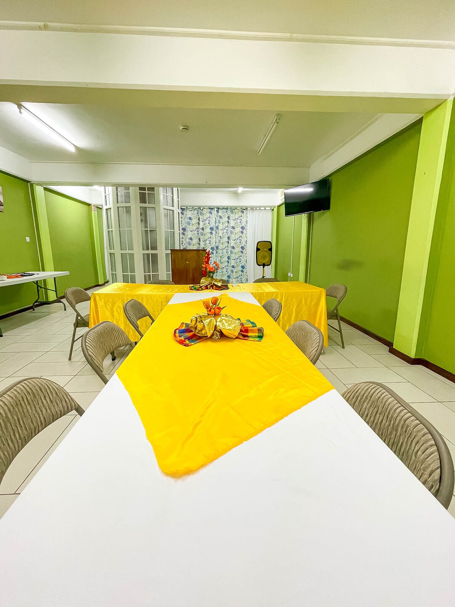 A conference room with a long table covered in yellow, surrounded by chairs, a speaker, and a window with curtains.