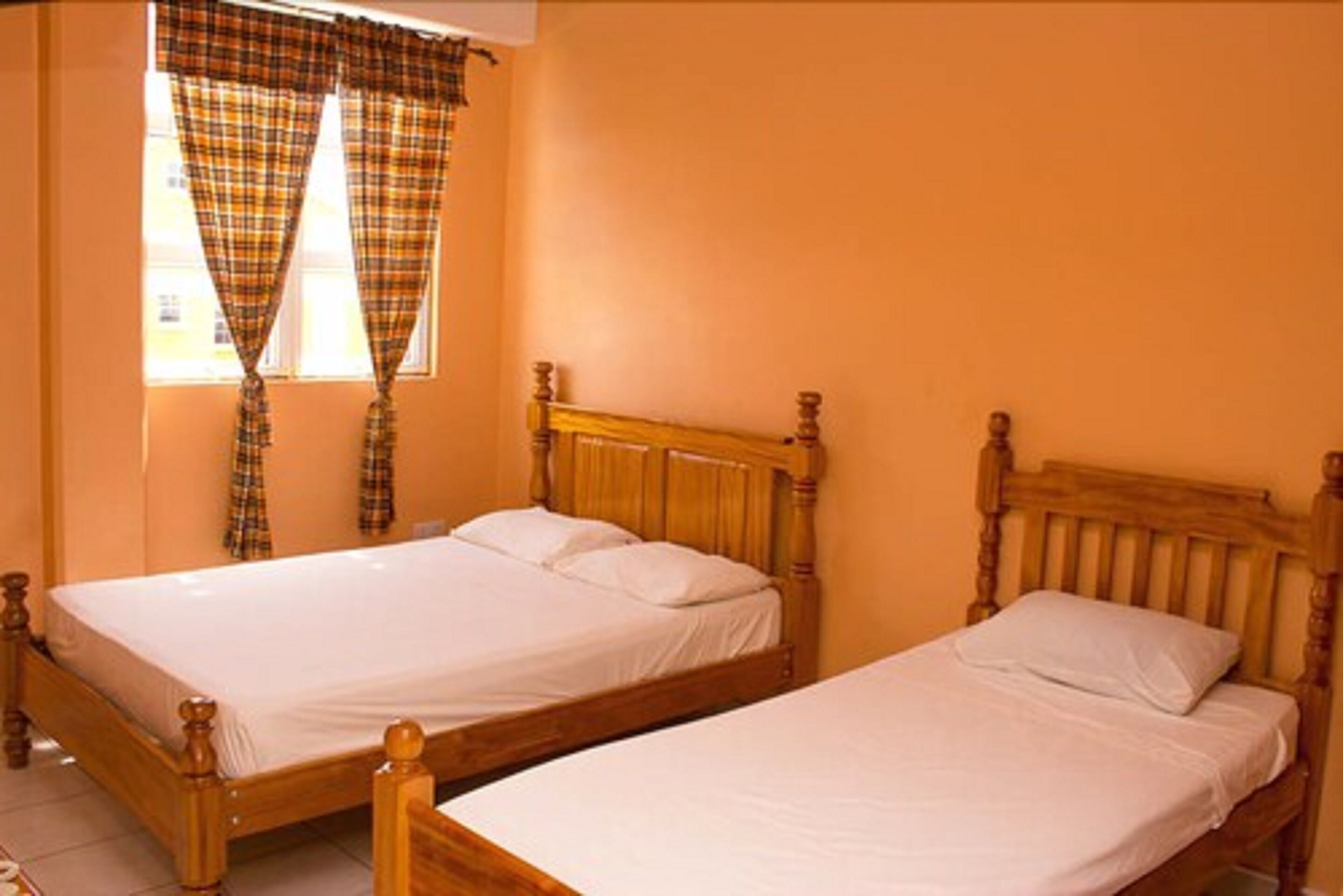 A room with two wooden beds, a window with plaid curtains, and a plain orange wall.