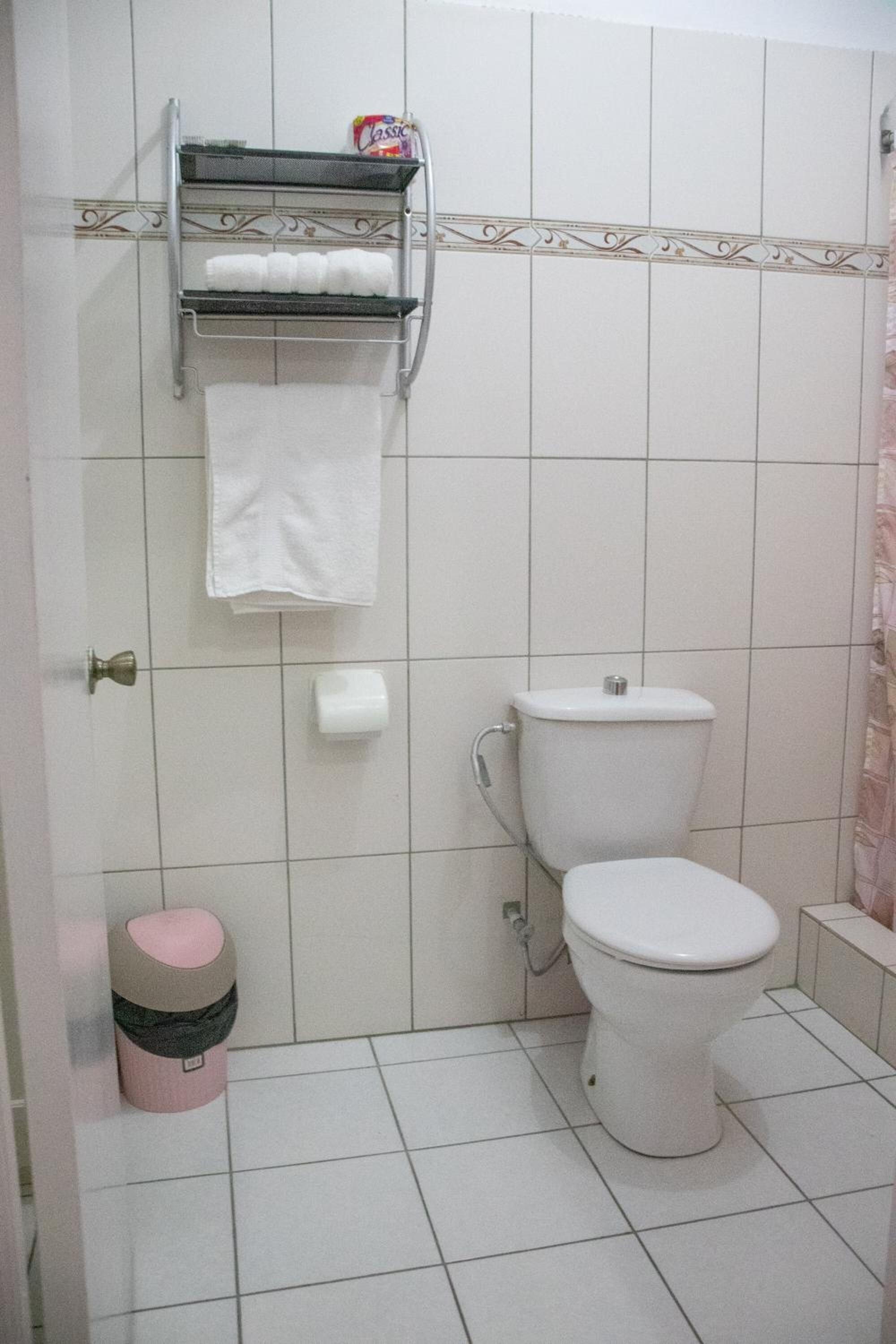 A bathroom with a toilet, a shelf with toiletries, a roll of toilet paper, and a trash bin.