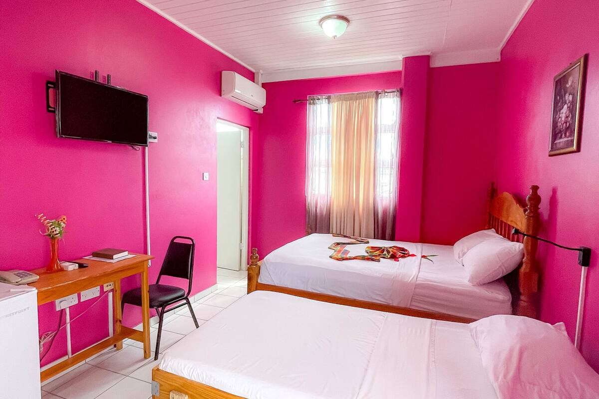 Una habitación con paredes rosas, una cama, un escritorio con silla, un televisor y un equipo de aire acondicionado.
