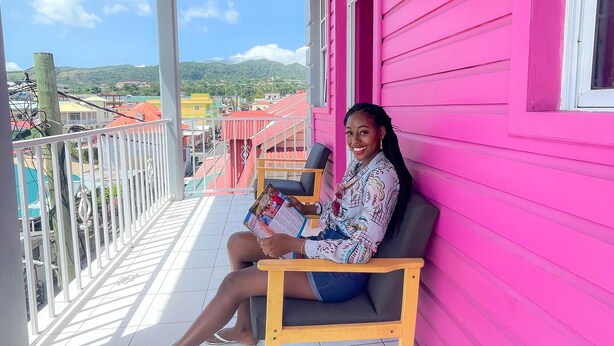 Une personne est assise sur un banc, sur un balcon, avec un bâtiment rose en arrière-plan.
