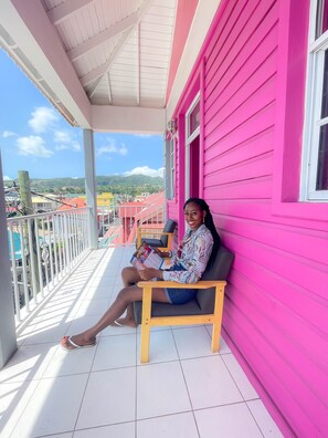 A person sitting on a bench on a balcony with a pink building in the background. - La Flamboyant Hotel (Roseau)