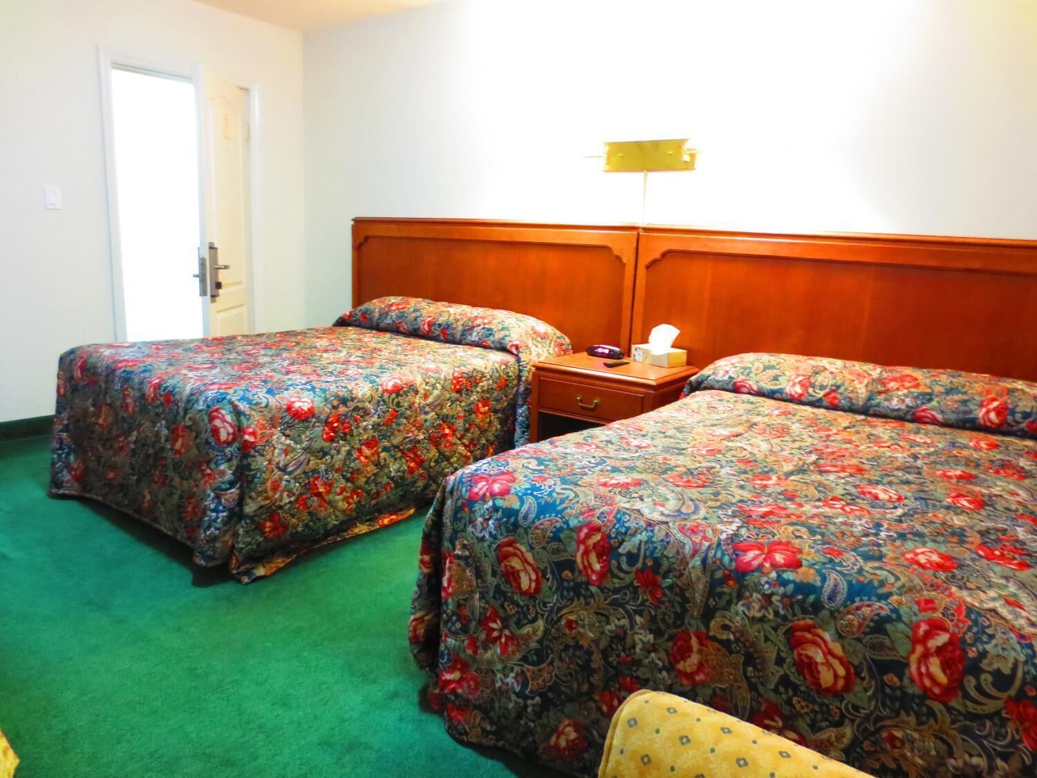 A hotel room with two beds, a wooden headboard, a bedside table with a lamp, and a green carpet.