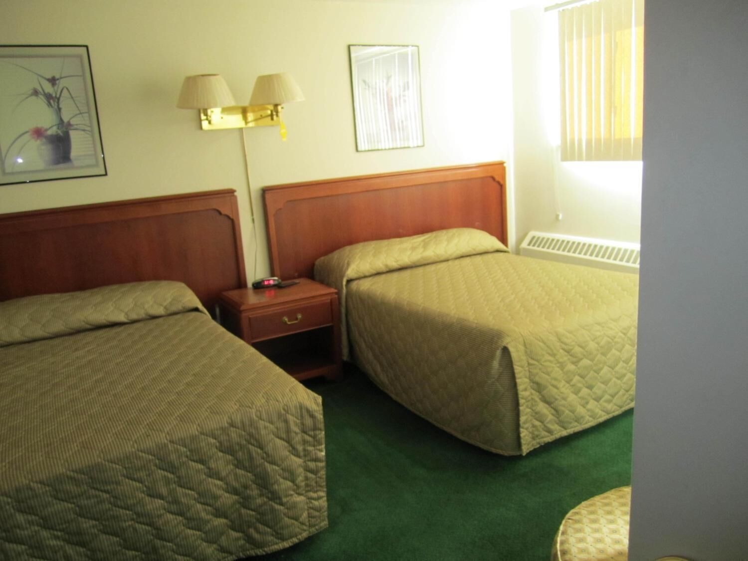 Une chambre d’hôtel avec deux lits simples, une table de chevet avec une lampe, un tableau encadré au mur et une fenêtre avec des stores.