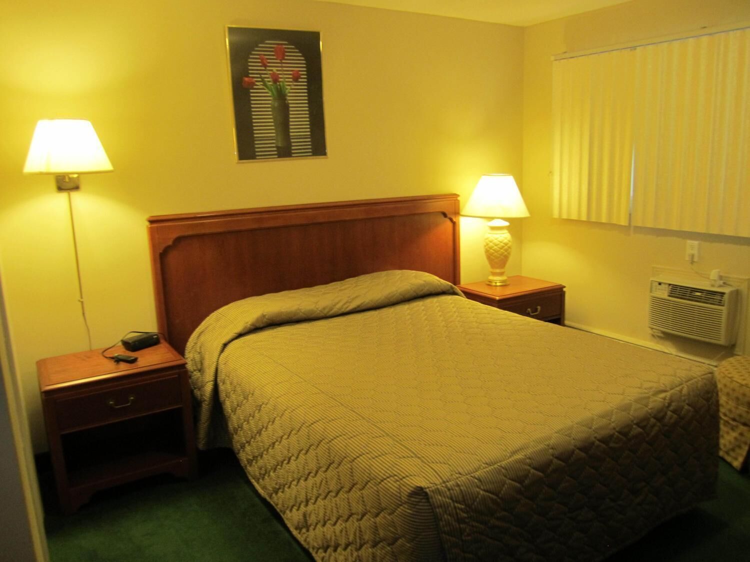 Una habitación de hotel con una cama, dos mesitas de noche, una lámpara sobre cada una, un aire acondicionado de pared y un cuadro enmarcado encima de la cama.
