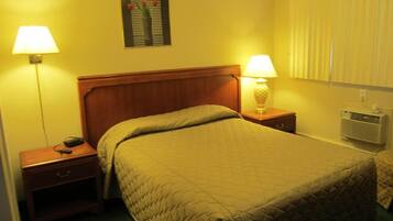 Una habitación de hotel con una cama, dos mesitas de noche, una lámpara sobre cada una, un aire acondicionado de pared y un cuadro enmarcado encima de la cama.