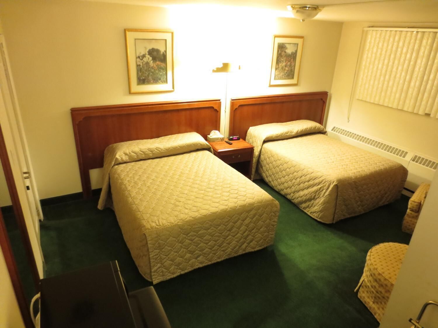 A hotel room with two single beds, a wooden headboard, a bedside table with a lamp, and two framed pictures on the wall.