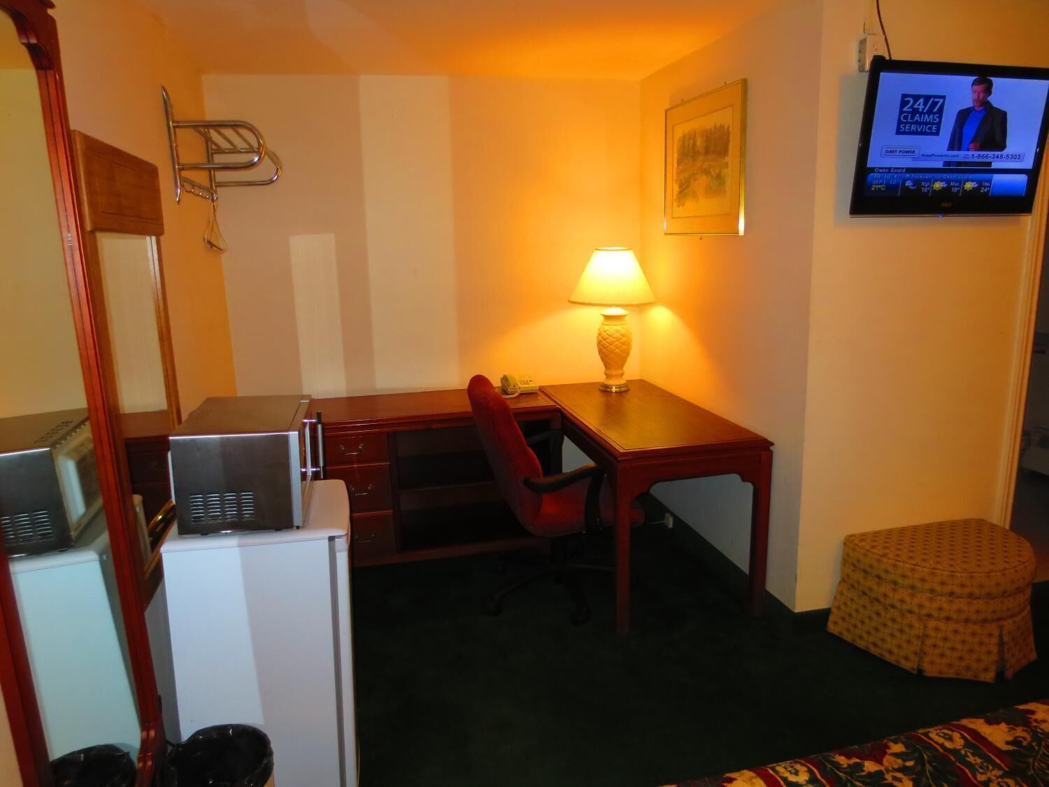 A hotel room with a desk, chair, lamp, television, and a small ottoman.
