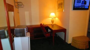A hotel room with a desk, chair, lamp, television, and a small ottoman.