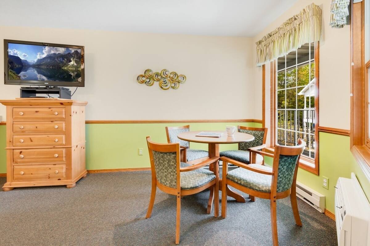 A room with a wooden dresser, a television, a round table with chairs, and a window with curtains.