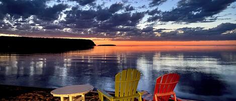 Escena de playa con tres sillas Adirondack y una pequeña mesa sobre la arena, con vista a una masa de agua tranquila y un atardecer de fondo.