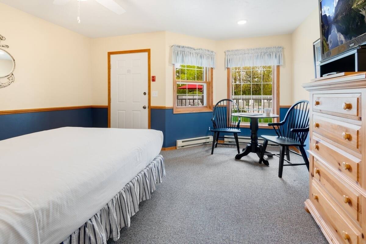 A bedroom with a bed, a dresser, a TV, a table with chairs, and windows with curtains.