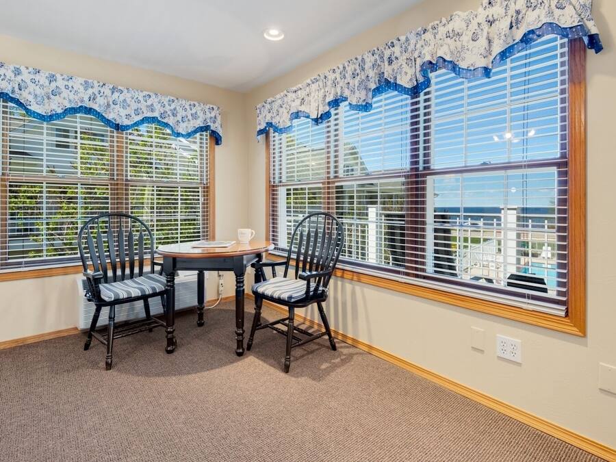 A room with two large windows, a table with chairs, and a view of a residential area.