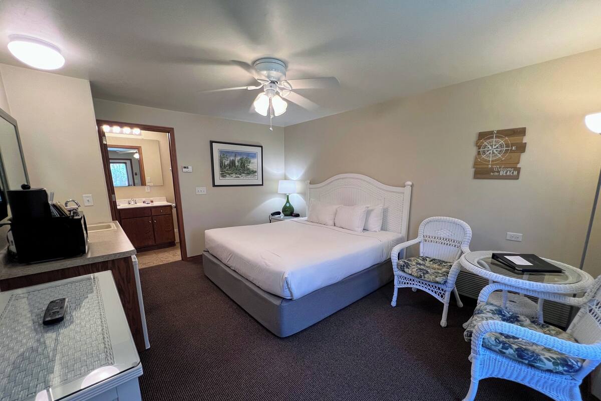A hotel room with a bed, a desk with a computer, a chair, a small table, and a ceiling fan.