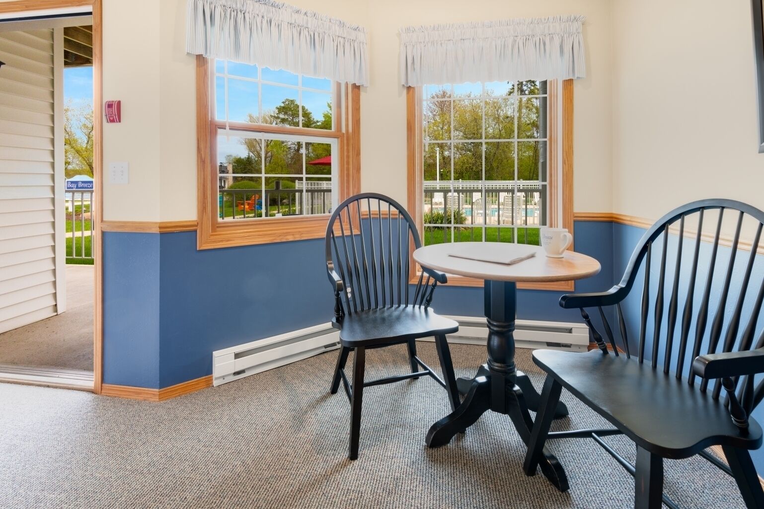A room with two black chairs around a small round table, a window with a view of greenery, and a door leading outside.