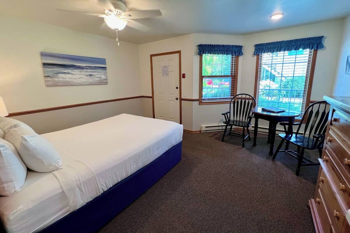 A hotel room with a bed, a desk, two chairs, a dresser, a ceiling fan, and a window with a view of the outside.