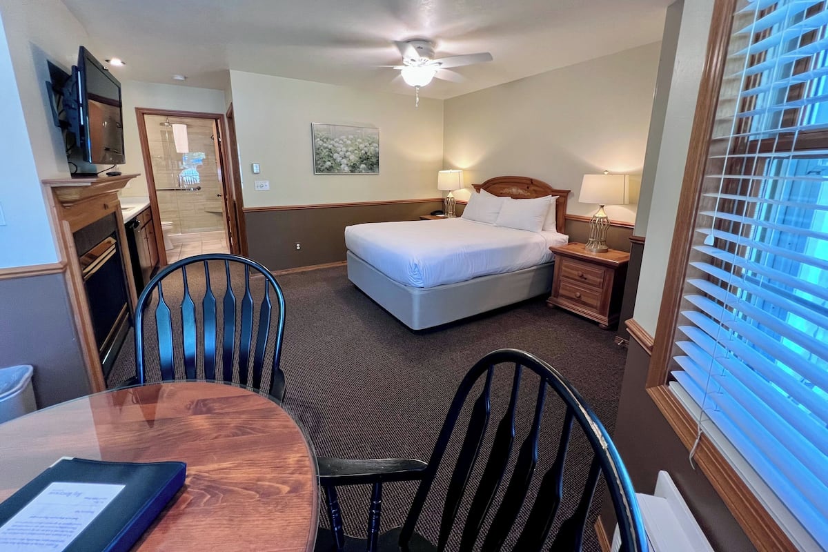 A hotel room with a bed, two bedside tables with lamps, a ceiling fan, a dining table with chairs, and a window with blinds.