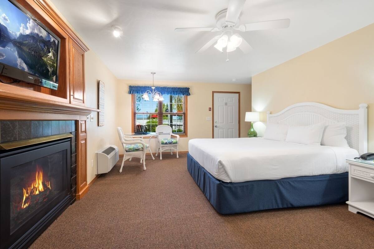 A hotel room with a fireplace, a bed, a desk with chairs, a television, and a ceiling fan.