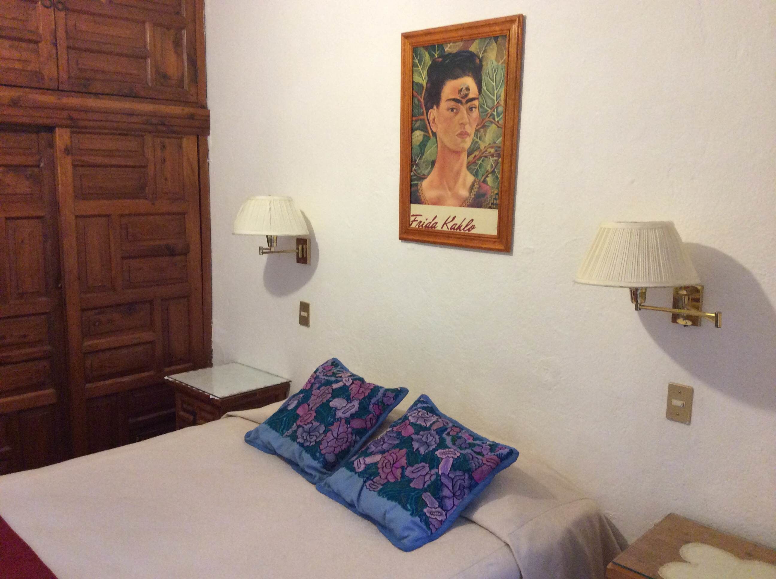 A bedroom with a bed, two pillows, a nightstand, a lamp, a wooden wardrobe, and a framed portrait on the wall.
