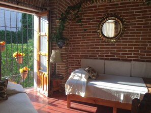 A room with a brick wall, a bed with a white bedspread, a wooden bench, a table with a lamp, and a window with hanging flower pots.