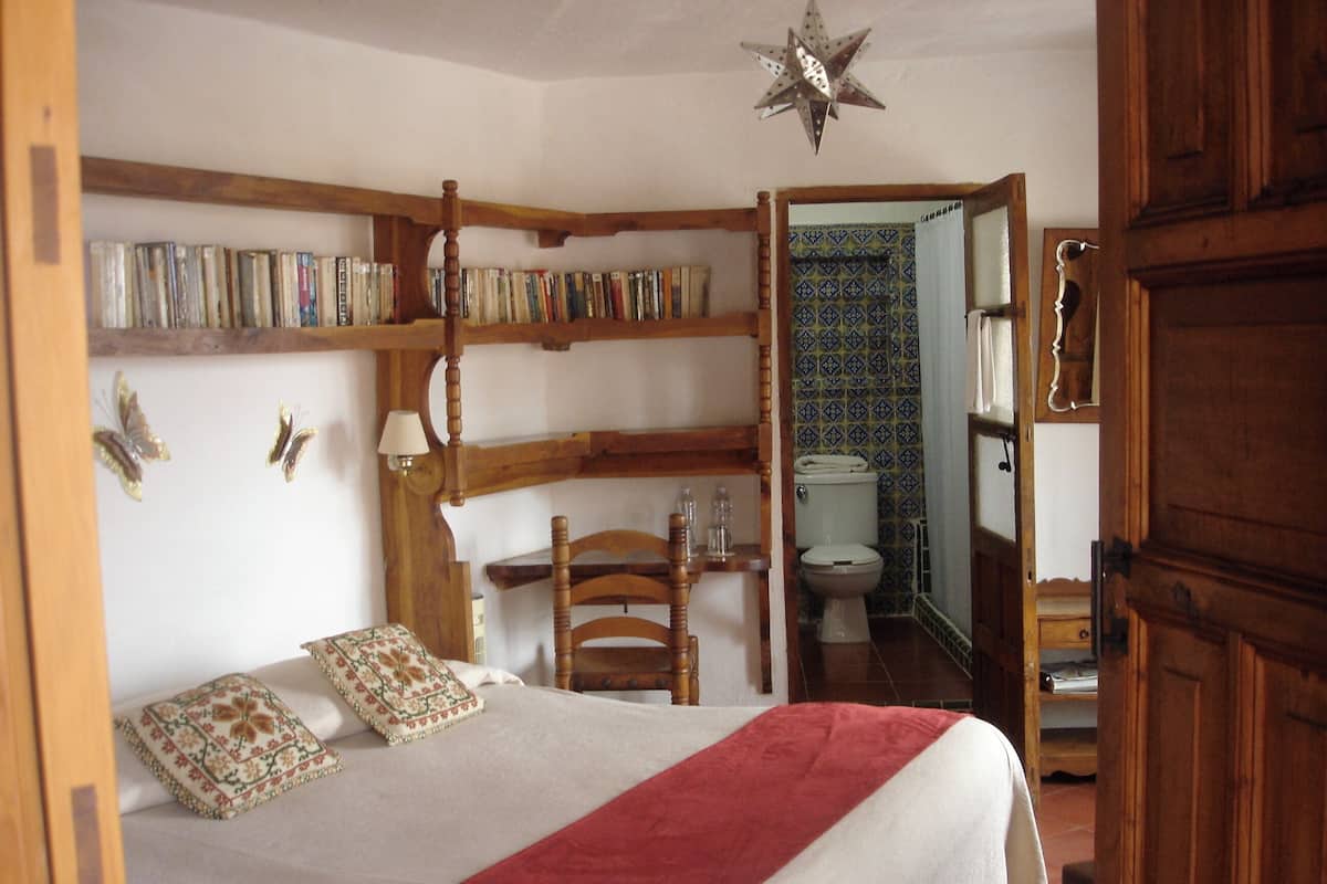 Une chambre à coucher avec un lit, une chaise en bois, une bibliothèque et une salle de bain visible à travers une porte ouverte.