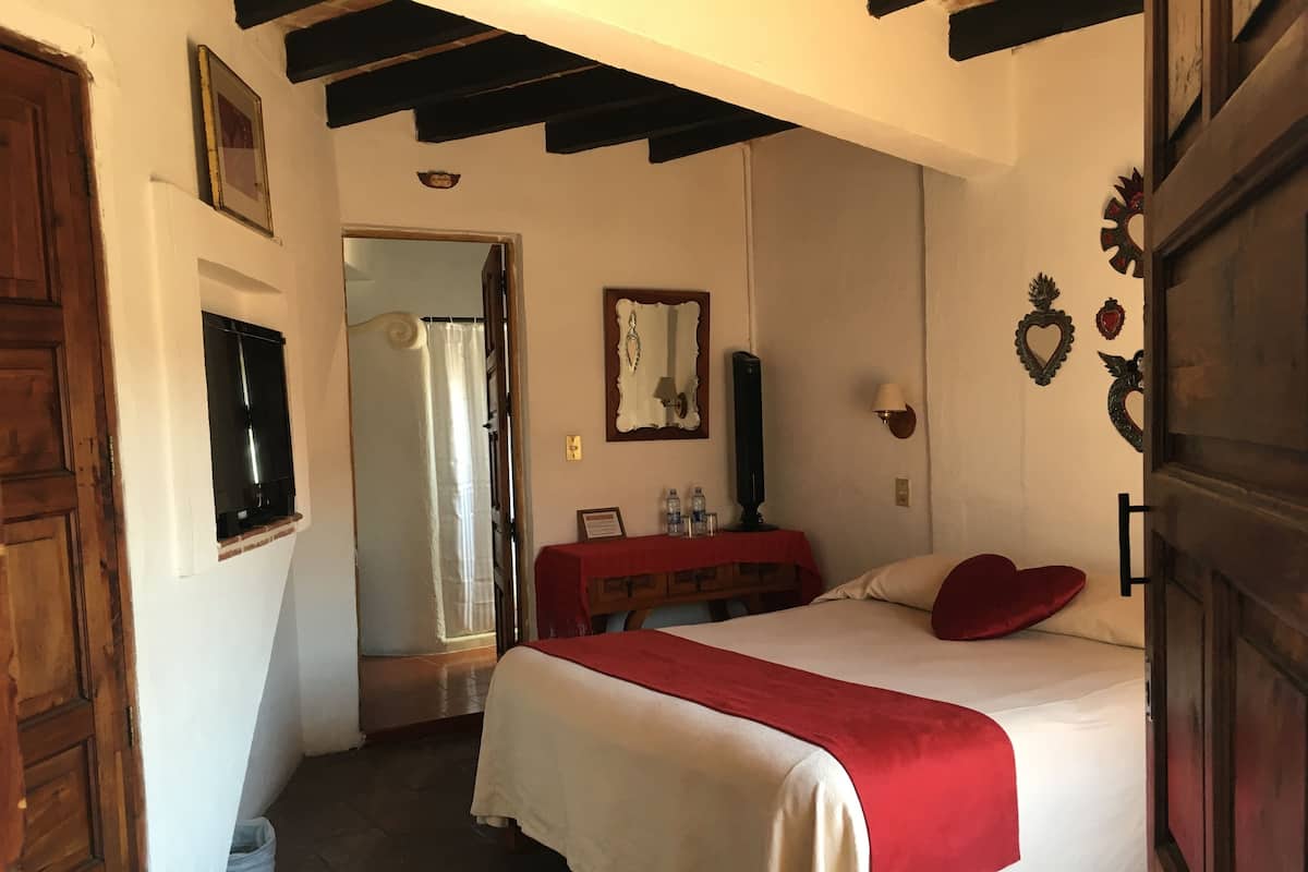 Une chambre à coucher avec un lit, une porte en bois, un miroir, une petite table recouverte d’une nappe rouge et un mur orné d’objets décoratifs.
