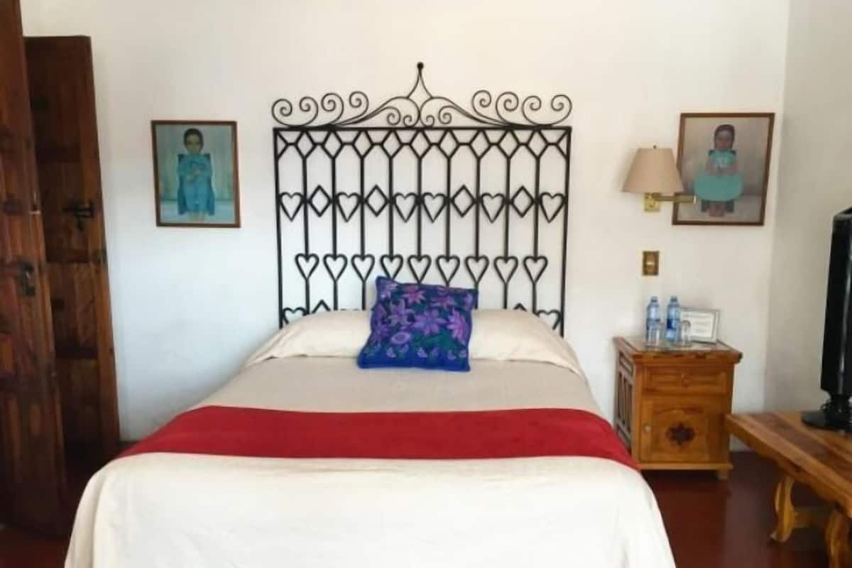Un lit avec une tête de lit ornée d’un motif en forme de cœur, une couverture rouge et un oreiller violet. Il y a deux tableaux encadrés au mur, une table de chevet en bois avec une lampe et un banc en bois.