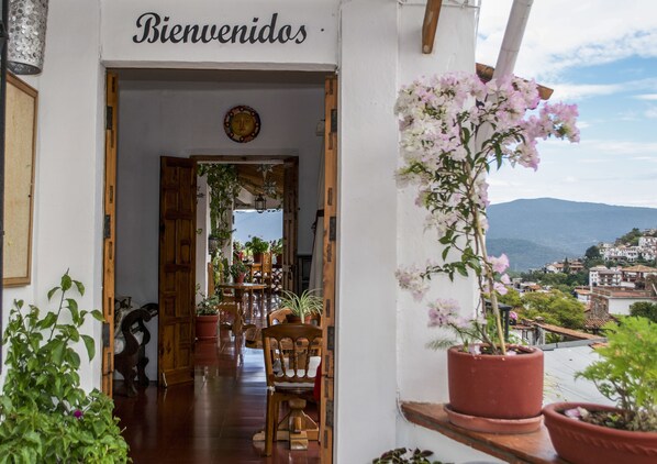 A welcoming entrance with a 'Bienvenidos' sign, wooden furniture, potted plants, and a view of a hillside town.