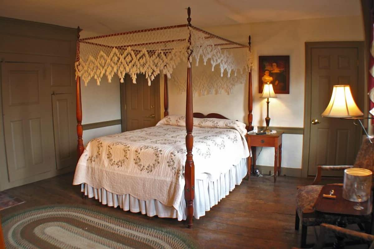 Een hemelbed met een bloemenbedekking, een nachtkastje met een lamp, een stoel en een deur in een kamer met houten vloeren en een kamerbreed tapijt.