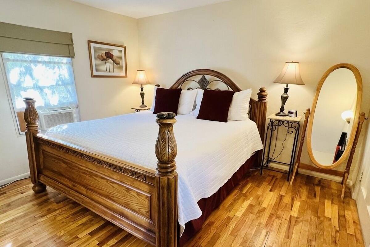 Una cama bien tendida con cabecera de madera, dos mesitas de noche con lámparas, un espejo grande y un cuadro enmarcado en la pared.