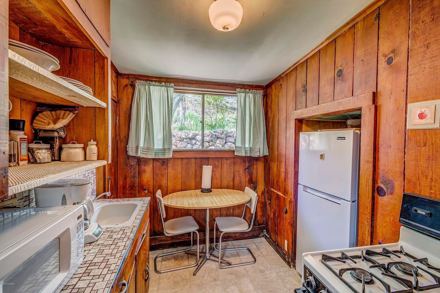 Une cuisine de taille réduite avec des murs en bois, une table à manger avec des chaises, un réfrigérateur, une cuisinière et une fenêtre avec des rideaux.