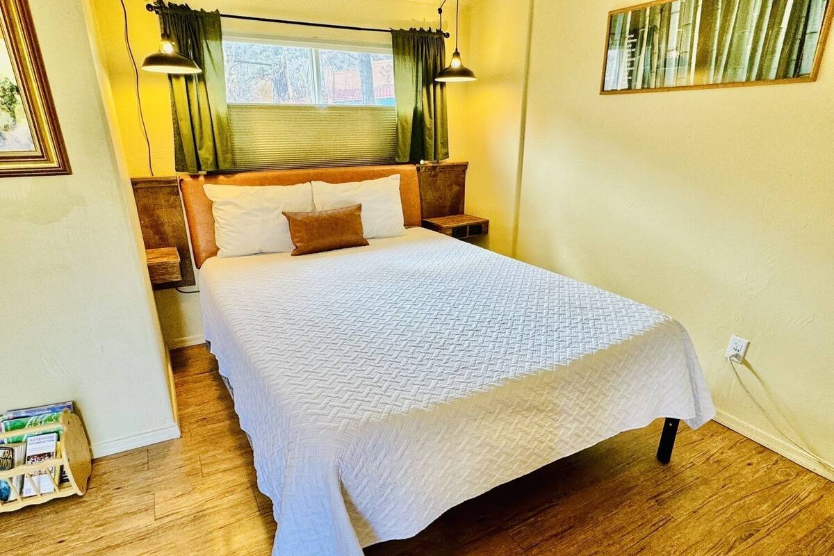 Una cama bien tendida en una habitación con suelo de madera, una ventana con cortinas y una lámpara de pared.