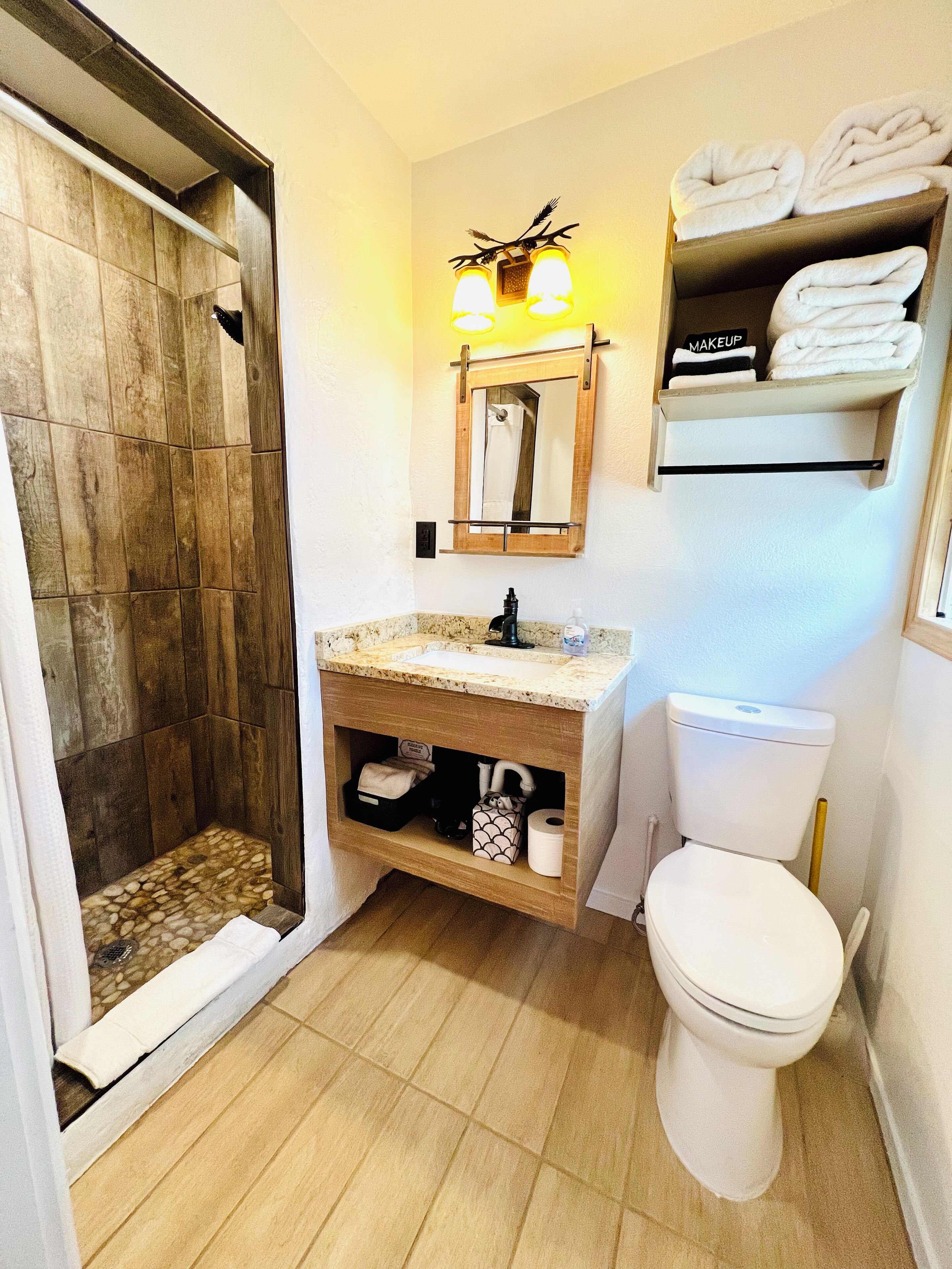 A bathroom with a wooden shower enclosure, a sink with a vanity, a toilet, and a shelf with towels.