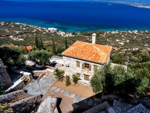 A stone building with a tiled roof, surrounded by greenery and overlooking a coastal area with houses and the sea. - Diotima's Homes (Kalamata)