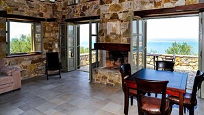 A stone-walled room with a fireplace, a dining table, and chairs. There are large windows offering a view of the sea. - Diotima's Homes (Kalamata)