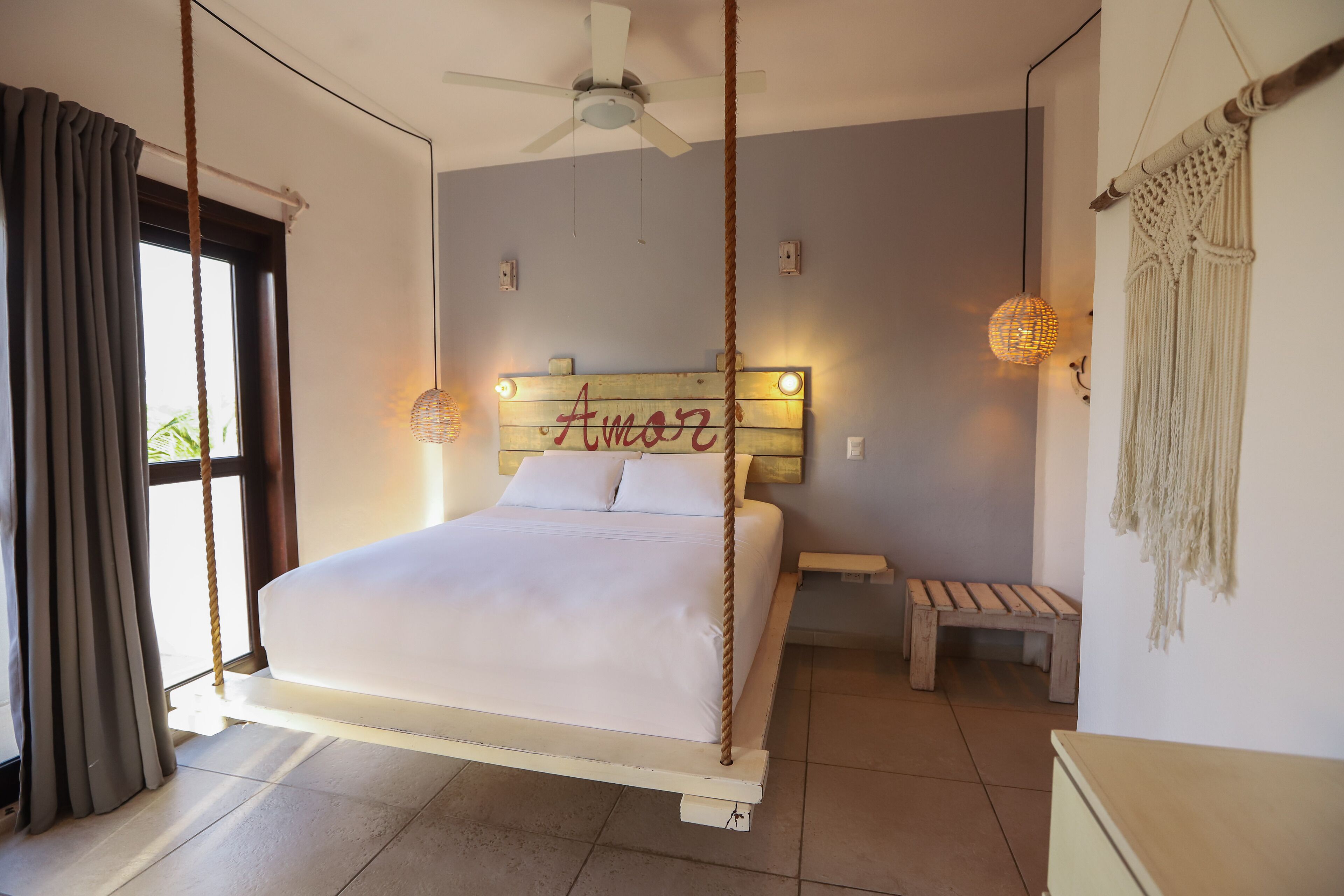 Un dormitorio con una cama suspendida del techo, una mesita de noche, un banco y un tapiz de macramé en la pared.