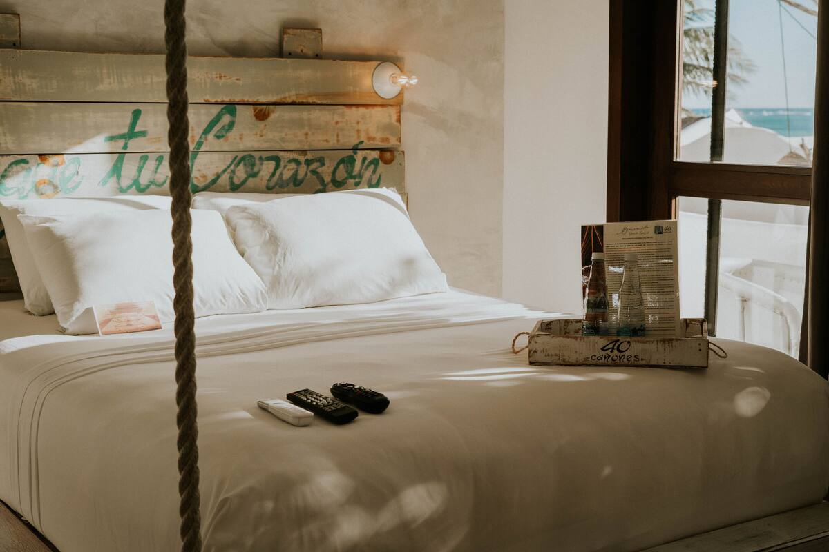 A bed with white linens, two remote controls, a menu, and a wooden headboard with a sign.
