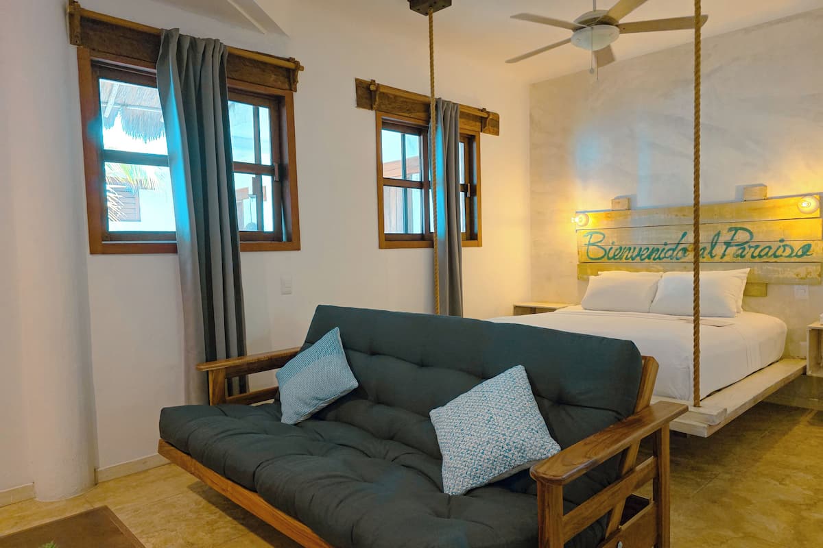 A room with a dark green sofa, two pillows, a bed with white bedding, a ceiling fan, and a sign reading "Bienvenidos al Paraíso".