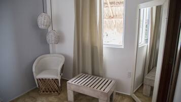 A room with a white chair, a wooden bench, a hanging light fixture, a window with a view of a straw structure, and a mirror reflecting part of the room.