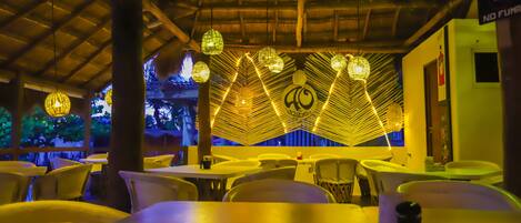 Un restaurante con mesas y sillas, iluminación decorativa y un techo de paja.