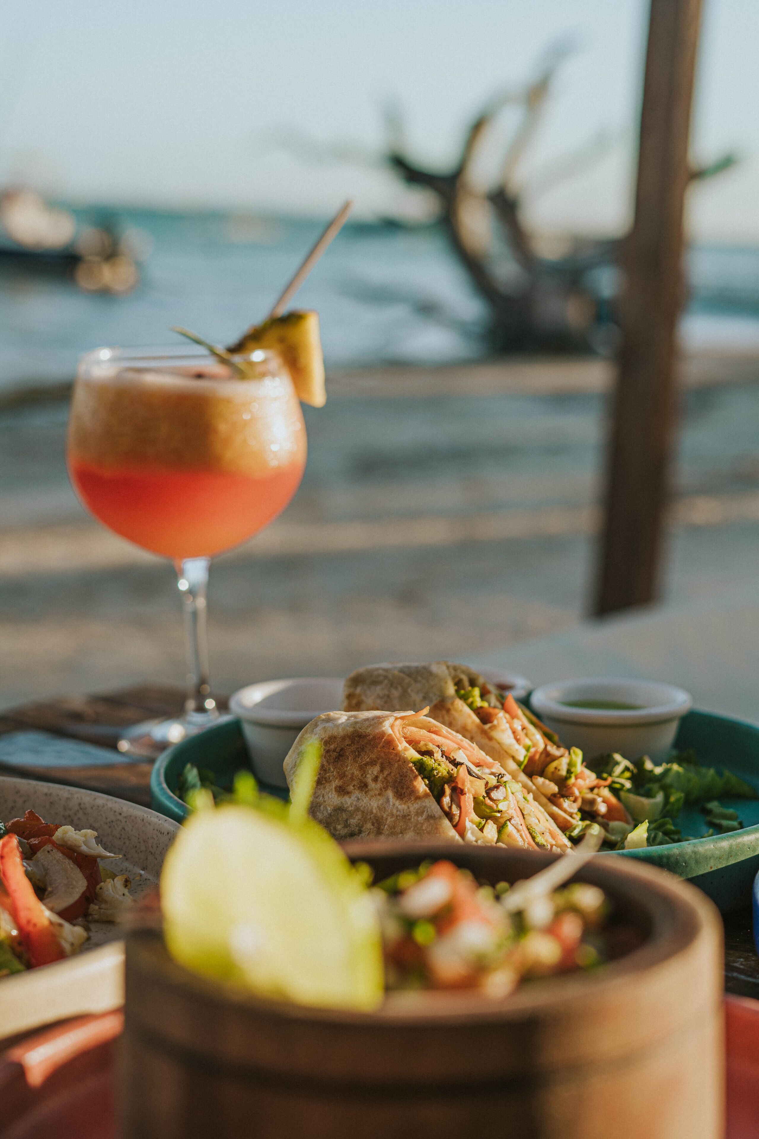 A table with a drink, sandwich, and salad, set against a coastal backdrop.