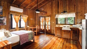 A wooden cabin with a bed, a dining table, a kitchen area with a stove, and a ceiling fan. - Posada Arco Iris (Roatan)