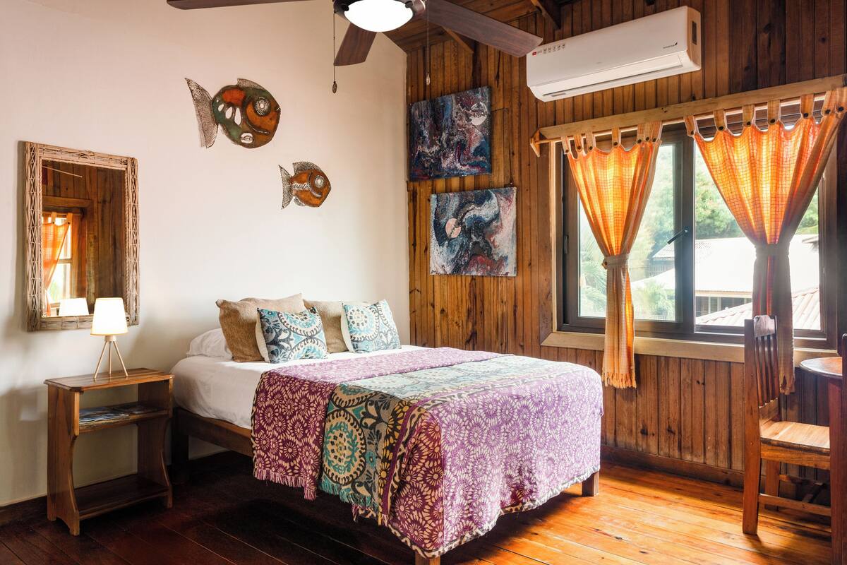 A wooden room with a bed, a nightstand, a ceiling fan, and a window with curtains.