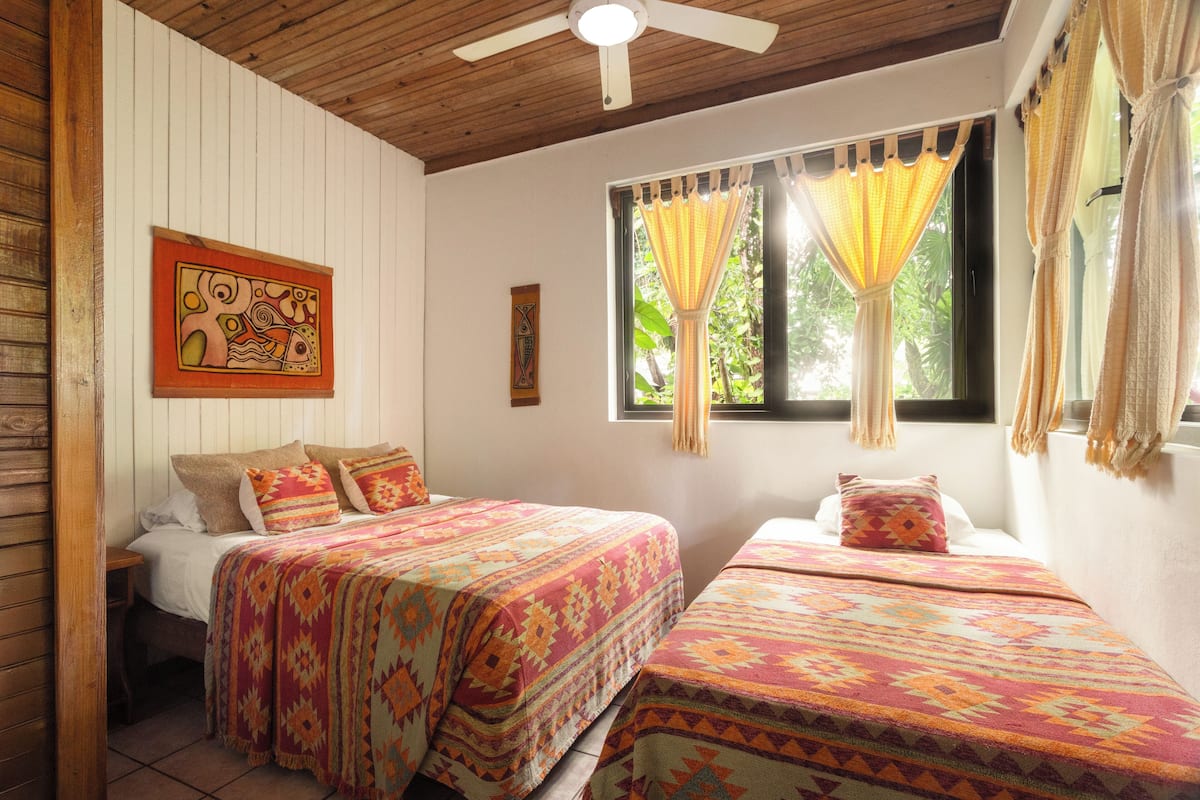 A room with two beds, a ceiling fan, a window with curtains, and a painting on the wall.