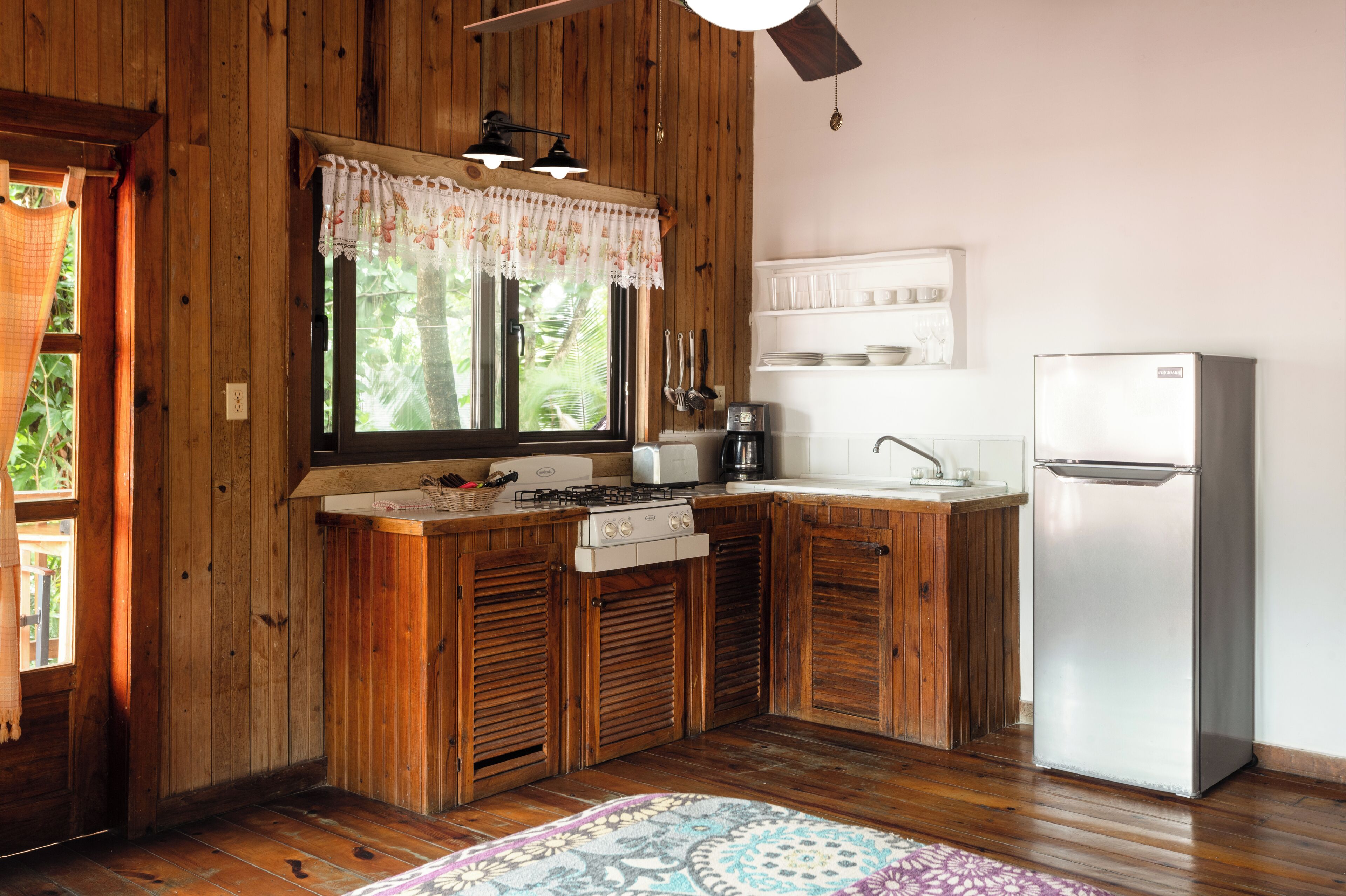 Una cocina compacta con armarios de madera, estufa a gas, refrigerador y ventana con cortina estampada.